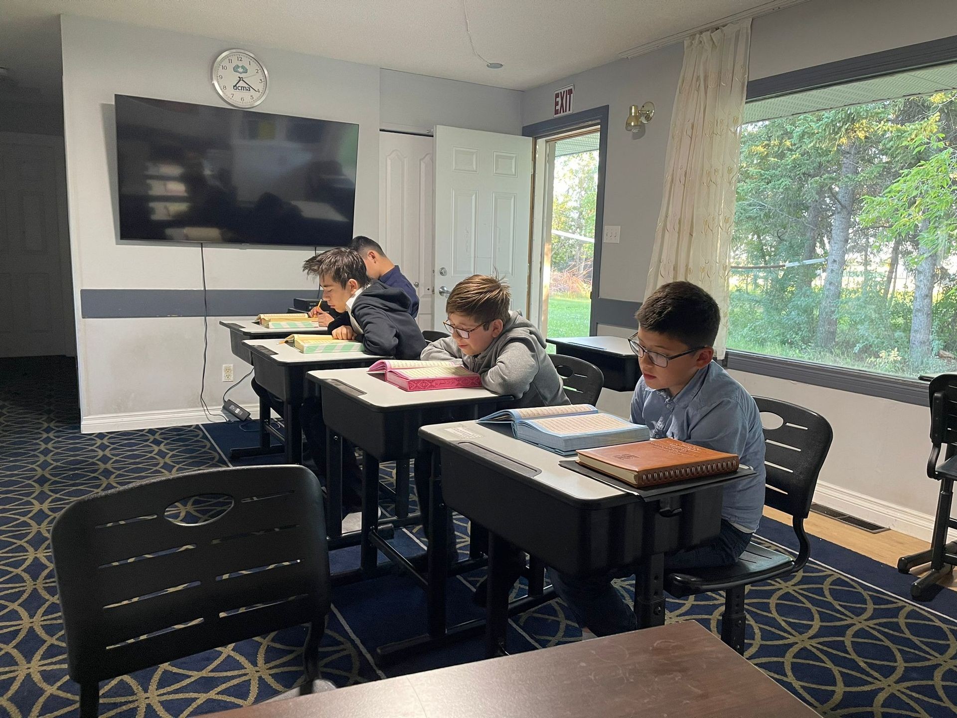 Children studying at desks in a classroom with a wall clock and large window view of trees outside.