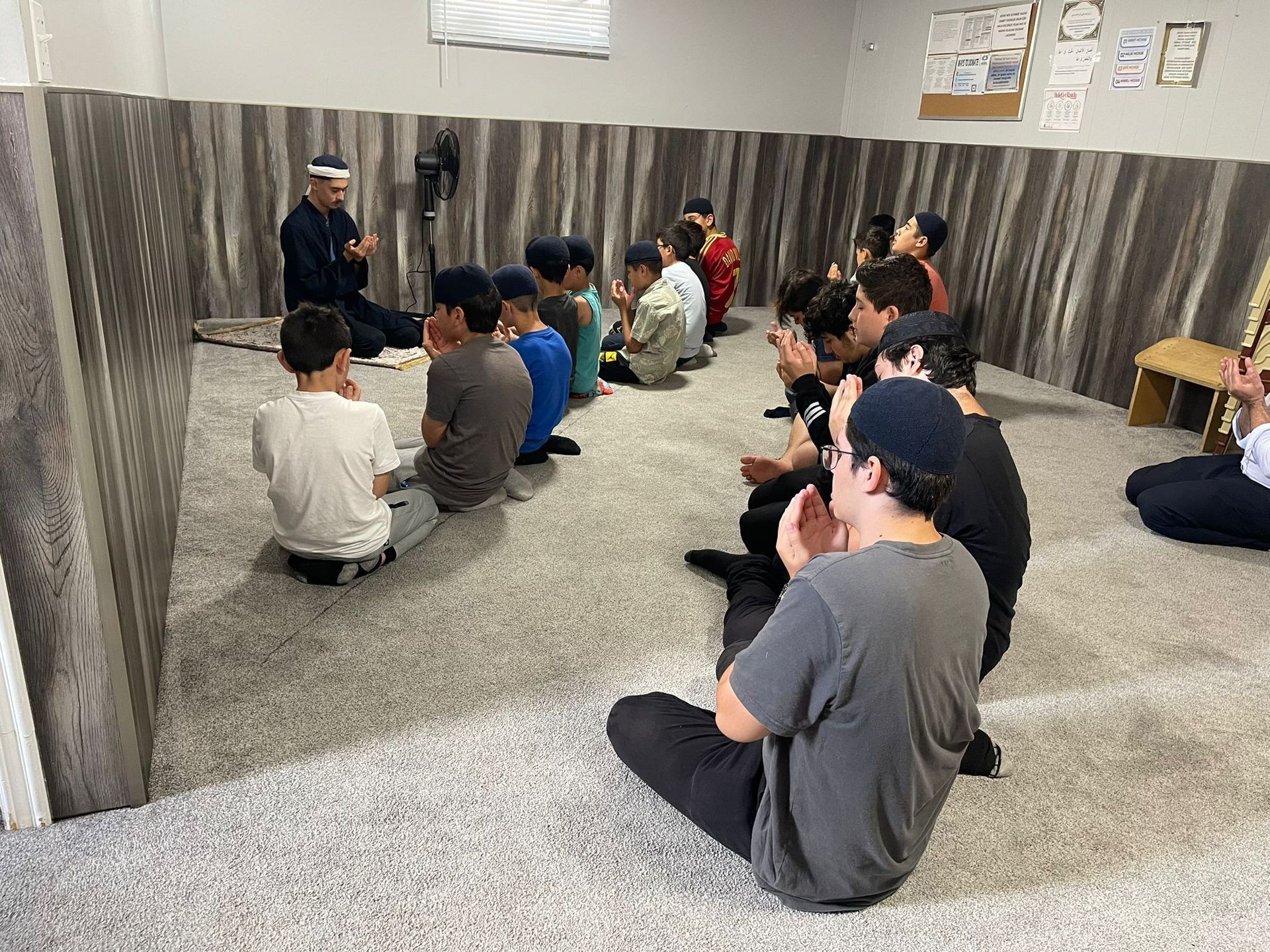 Group of people sitting on the floor in prayer, inside a room with wooden panels and a fan.