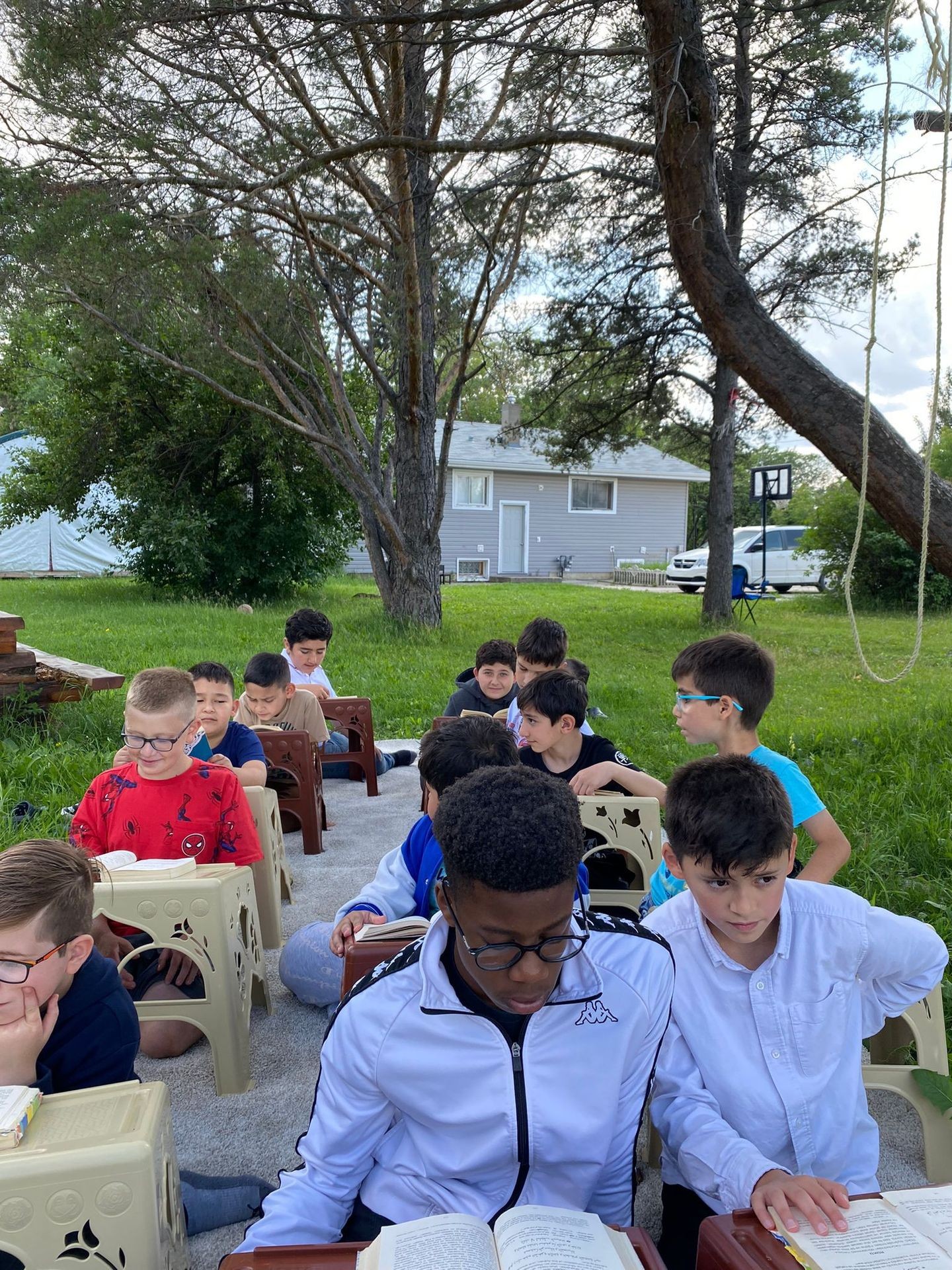 Group of kids sitting outdoors on stools reading books under a tree with a house in the background.