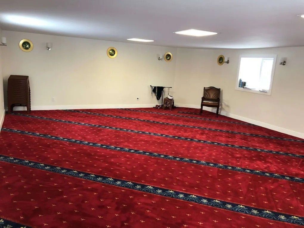 Spacious room with red carpet and decorative patterns, featuring a few chairs and wall hangings.